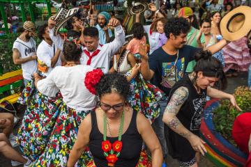 A group of people in colorful clothing dance. A woman is centered with a black tank top and a heavily beaded floral necklace of red, yellow, and orange. Red flowers sit on the left of her curls and she smiles at the ground.