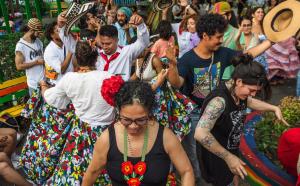 A group of people in colorful clothing dance. A woman is centered with a black tank top and a heavily beaded floral necklace of red, yellow, and orange. Red flowers sit on the left of her curls and she smiles at the ground.